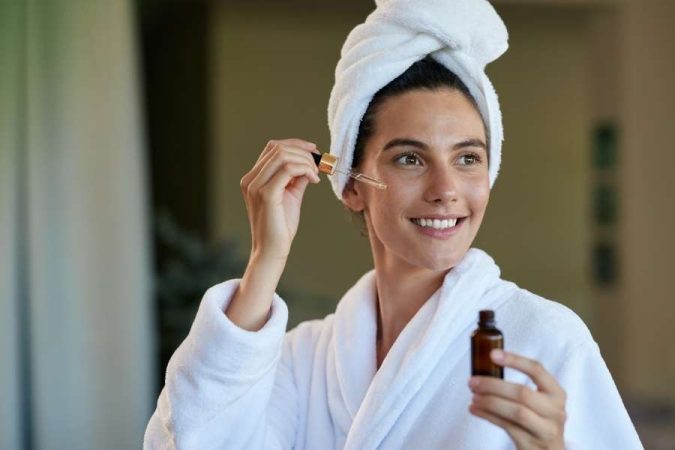 Une jeune femme en peignoir et serviette sur la tête applique des huiles végétales pures sur son visage avec une pipette.